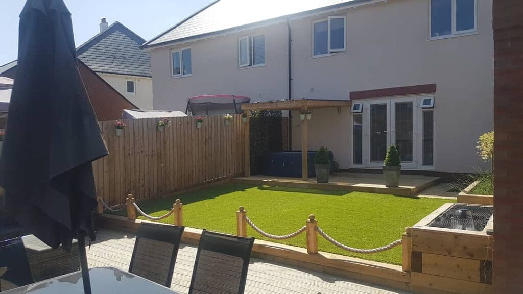 A backyard garden with artificial grass, a wooden fence, and a small patio area. There's a covered seating area, a trampoline, and potted plants. A rope barrier separates the grass from the decked patio with outdoor furniture.