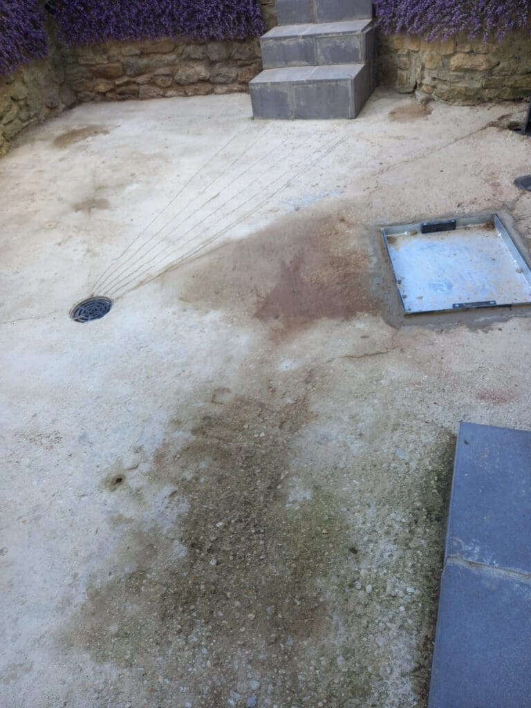 A concrete patio with a drain in the center and steps leading up. There are sprays of water trickling towards the drain. A metal tray is on the right side, and there are stone walls with purple plants on top.