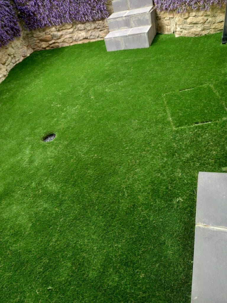 A small backyard with well-maintained artificial grass, bordered by stone walls. A few stone steps lead to a higher level, and the walls are adorned with purple flowering plants.