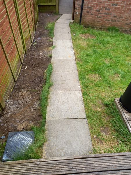 A narrow stone path runs through a small garden, bordered on one side by a red brick wall and on the other by a patch of grass. The path leads to a wooden gate.