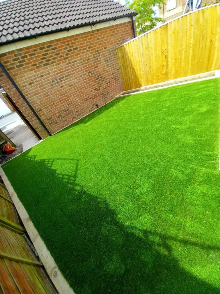 A small yard with bright green artificial grass next to a brick building and a wooden fence. The area is enclosed, and sunlight creates shadows on the grass.