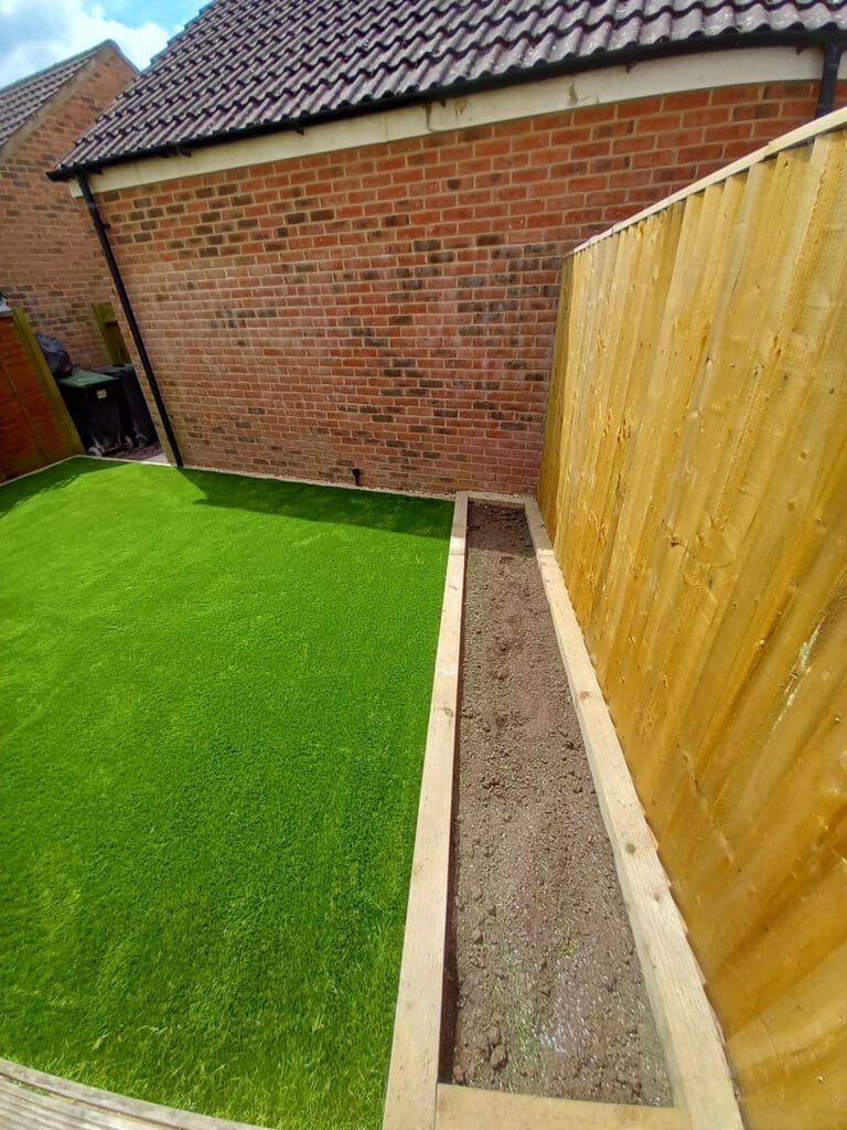 A small backyard with artificial grass bordered by red brick walls and a wooden fence. A narrow, empty flower bed runs along the right side, next to the fence.