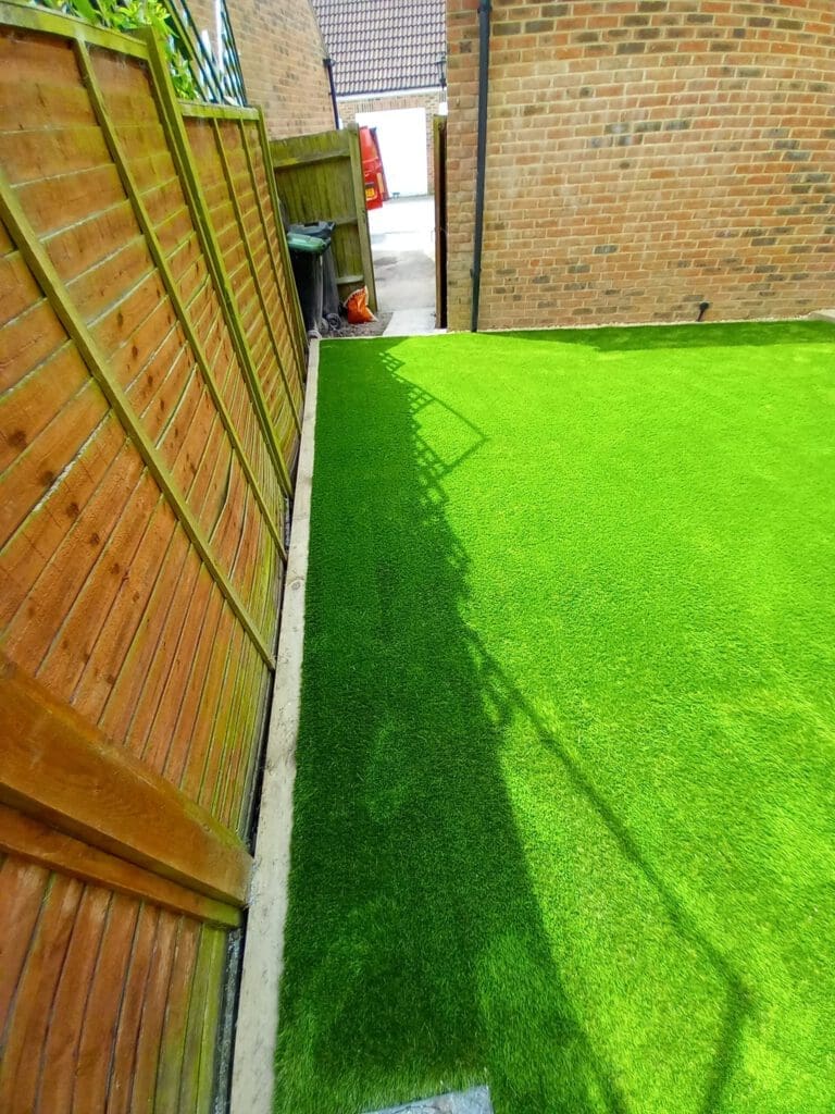 A garden area with bright green artificial grass bordered by a wooden fence on the left and brick buildings. A gate is open in the background, leading to a paved driveway.