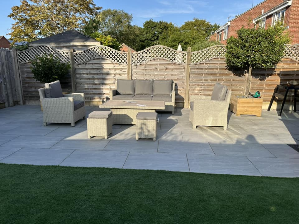 A patio with beige wicker furniture, including a sofa, two chairs, and two small tables. The area is paved with large tiles, bordered by a wooden fence and some trees. A patch of artificial grass is visible in the foreground.