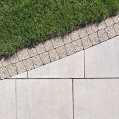 A neatly manicured lawn borders a path made of rectangular stone tiles. Separating the grass and the tiles is a strip of small, square stone pavers set at an angle, creating a visually pleasing geometric pattern.