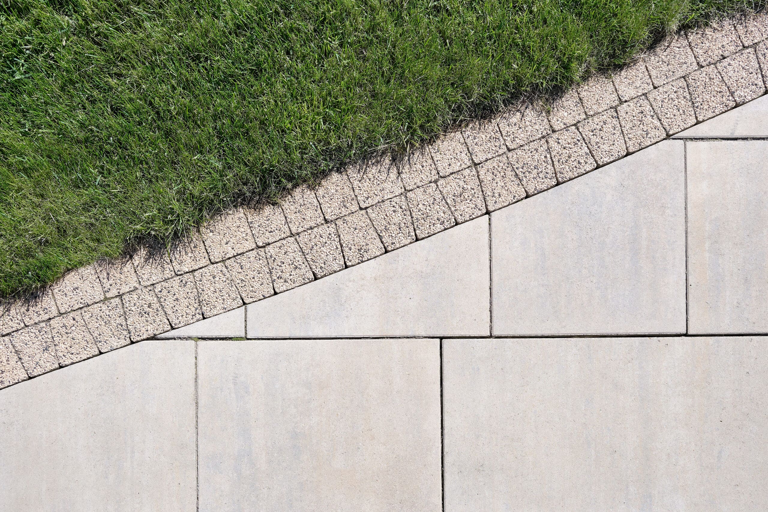 A neatly manicured lawn borders a path made of rectangular stone tiles. Separating the grass and the tiles is a strip of small, square stone pavers set at an angle, creating a visually pleasing geometric pattern.