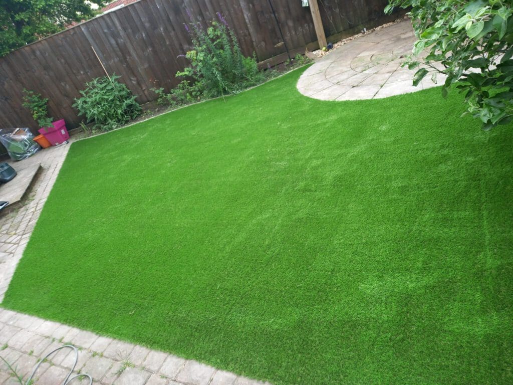 A backyard with a lush green lawn, bordered by a brown wooden fence. There are circular stone pathways in the corner and various plants growing along the fence. Several objects, including a pink container, are visible near the fence.