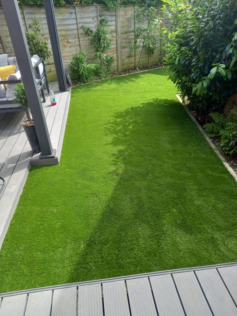 A small backyard features a neatly manicured rectangular lawn surrounded by shrubs and leafy plants. In the background, there's a wooden deck with outdoor furniture, including a gray sofa and a yellow pillow. A tall fence encloses the area.