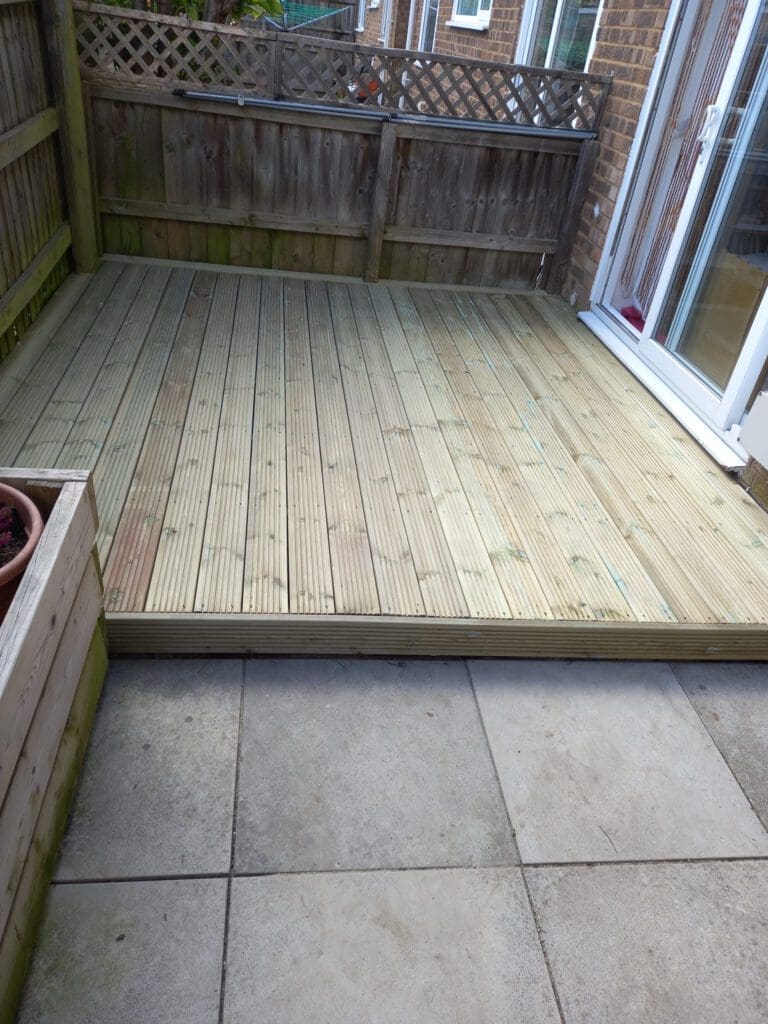 A wooden deck adjacent to a house with sliding glass doors, surrounded by a wooden fence. The deck is raised above a stone-tiled patio. A large planter is visible on the left side.