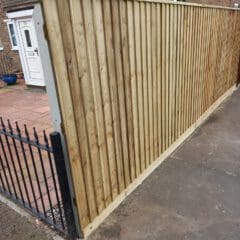 A tall wooden fence with vertical panels is seen along a sidewalk in front of a house. The house has a white door and brick walls. A black metal gate is adjacent to the fence.