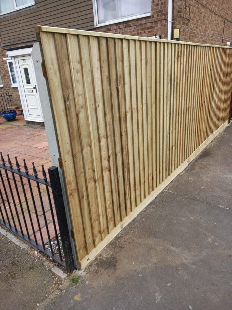 A tall wooden fence with vertical panels is seen along a sidewalk in front of a house. The house has a white door and brick walls. A black metal gate is adjacent to the fence.