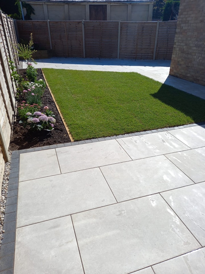 A backyard with a neatly mowed lawn bordered by a flower bed with pink and purple flowers. The foreground features a light gray stone patio, and a wooden fence surrounds the area. Sunlight casts shadows on the ground.