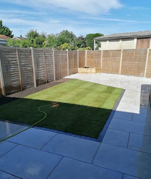 A small backyard features a neatly mowed grass area bordered by light gray stone tiles. Wooden fencing surrounds the yard. A garden hose is partially visible on the grass, and a clear blue sky is overhead.