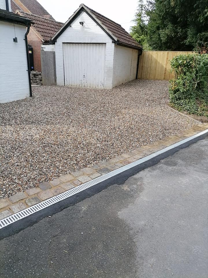 A gravel driveway leads to a white garage with a tiled roof. A wooden fence is on the right, and a house wall is on the left. A strip of new asphalt with drainage runs along the edge of the driveway.