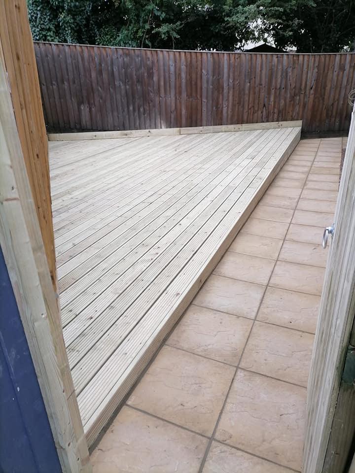 A garden area features a newly installed wooden deck next to a section of beige tiles. The space is enclosed by a brown wooden fence, and trees are visible in the background.