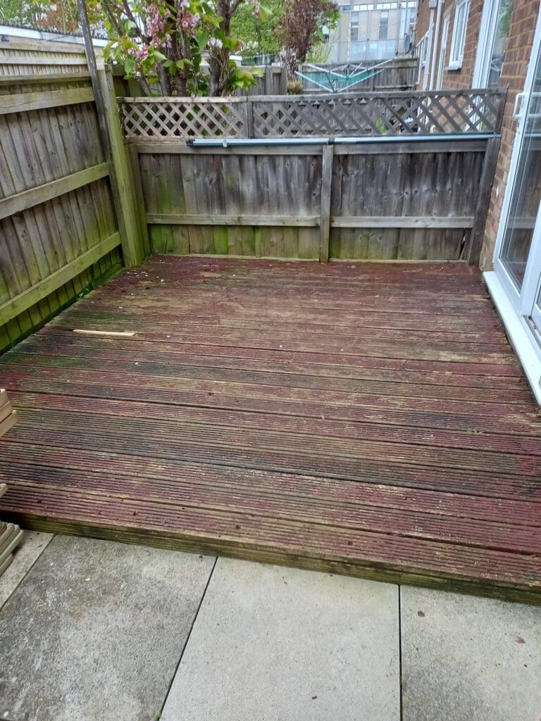 A wooden deck with visible wear and discoloration, surrounded by a weathered wooden fence. There are some trees in the background and a building visible behind the fence. The deck connects to a concrete patio area.