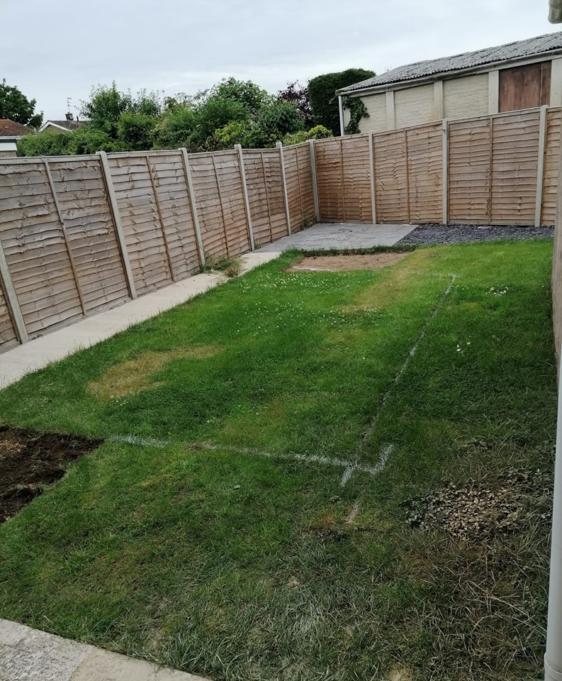 A backyard with a grass lawn surrounded by wooden fencing. The lawn has a few patches where grass is missing, and there are outlined rectangular areas on the grass. A small paved area is visible near the fence.