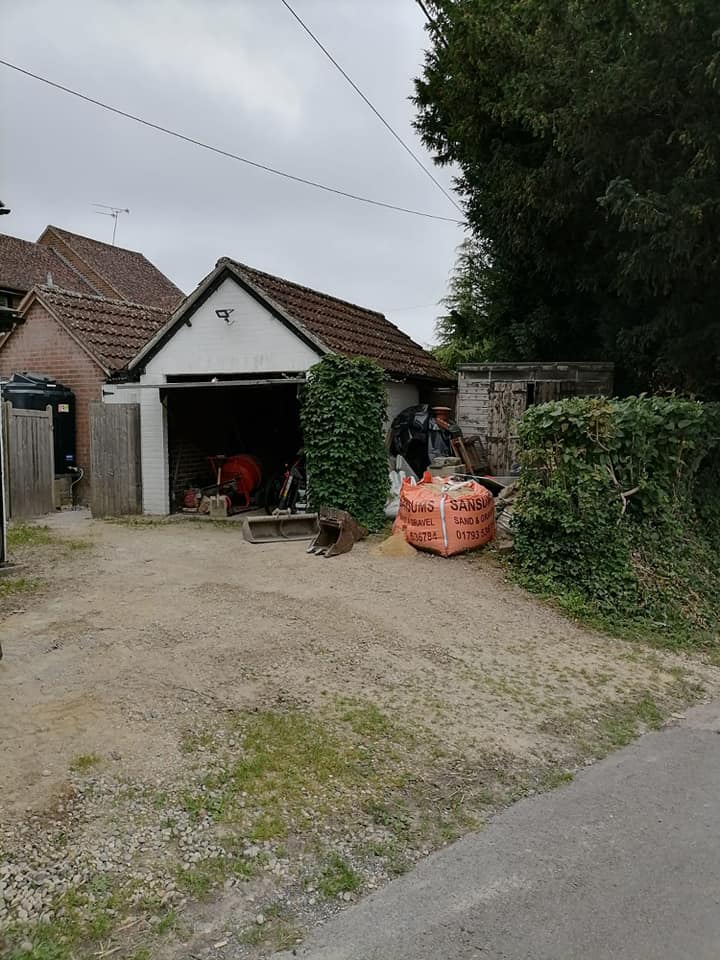 A small garage with a white facade and a red-tiled roof, partially hidden by ivy and shrubs. Two large orange bags are placed in front, with various tools and equipment scattered around. A clear sky is visible above.