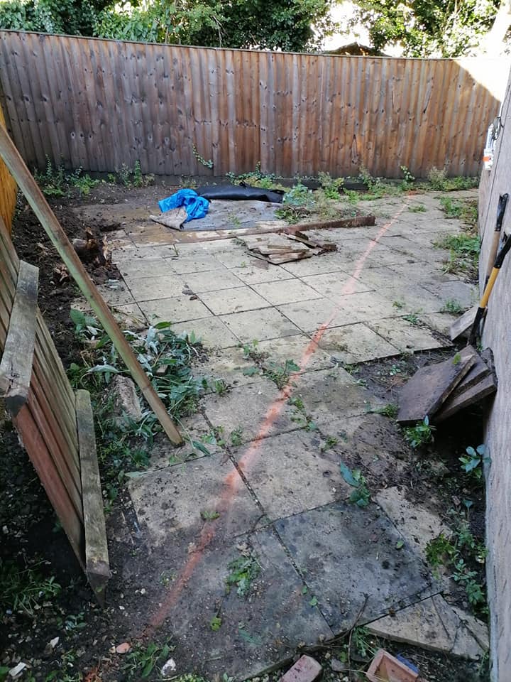 A backyard with a patio covered in large stone tiles. Weeds grow between the tiles, and a wooden fence borders the area. Some boards are broken or scattered, and a blue tarp is seen in the corner. A red chalk line marks part of the ground.