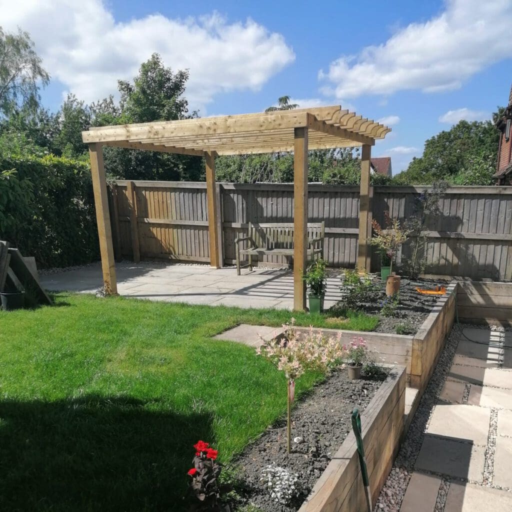 A backyard with a wooden pergola on a paved patio, surrounded by a wooden fence. The area includes a small garden bed with plants and flowers, and a grassy lawn. The sky is partly cloudy.