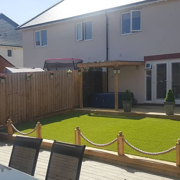 Backyard with artificial grass, a wooden pergola, potted plants, and a wooden fence. A glass table set is in the foreground, with a trampoline visible over the fence. Double doors lead into a two-story house with beige walls.