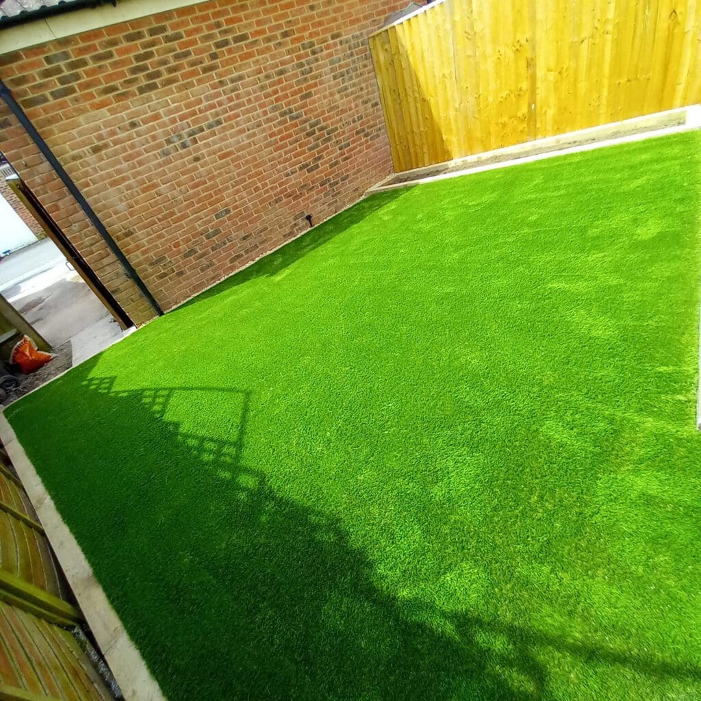 A small backyard with vibrant green artificial grass surrounded by a red brick wall and a wooden fence. Shadows of a metal fence are cast on the grass, and there's a glimpse of a concrete path and a red object in one corner.