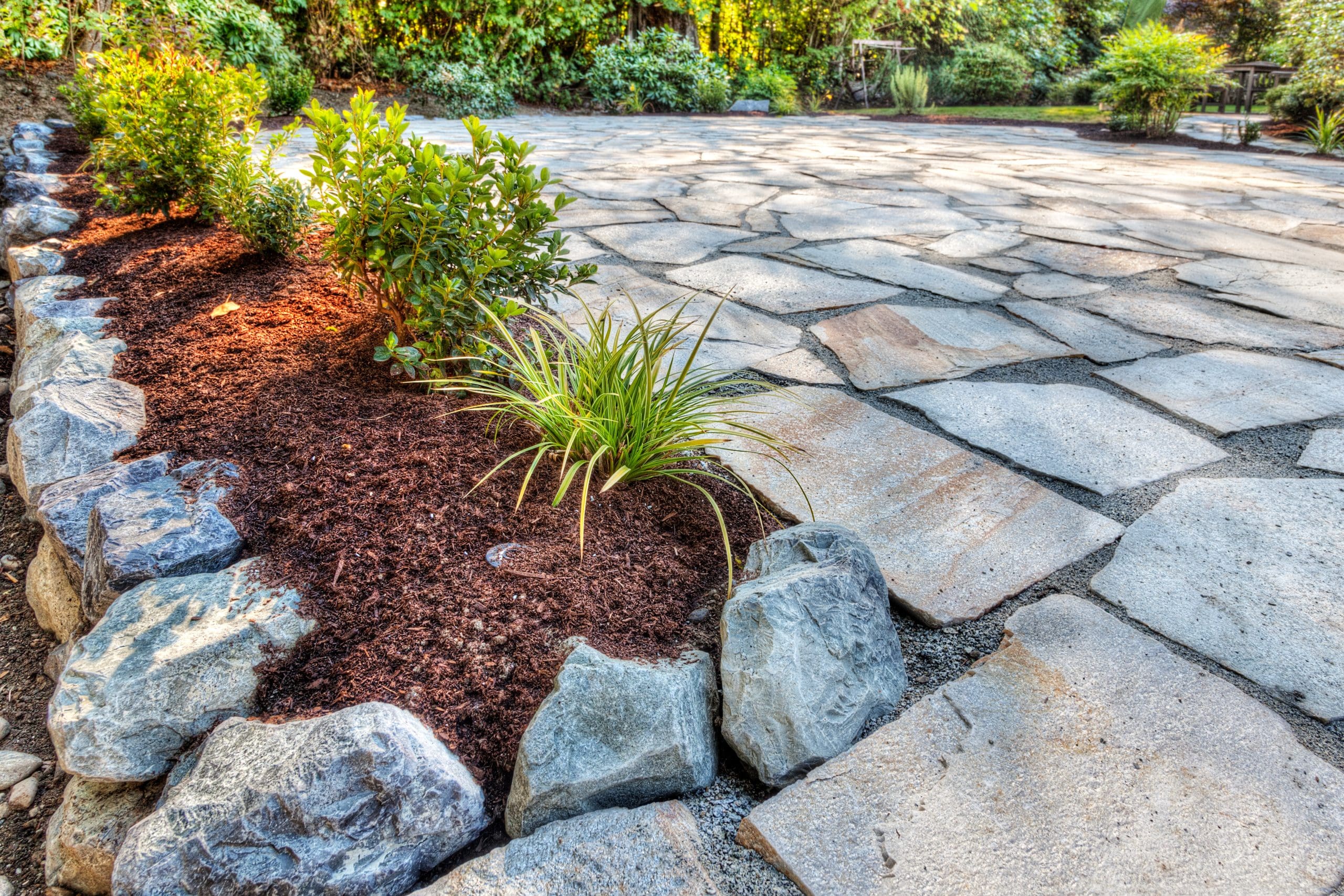 A landscaped garden path made of large, irregular grey stone slabs bordered by a flower bed with mulch and rocks. Green plants and small shrubs add color against the backdrop of lush trees and foliage.