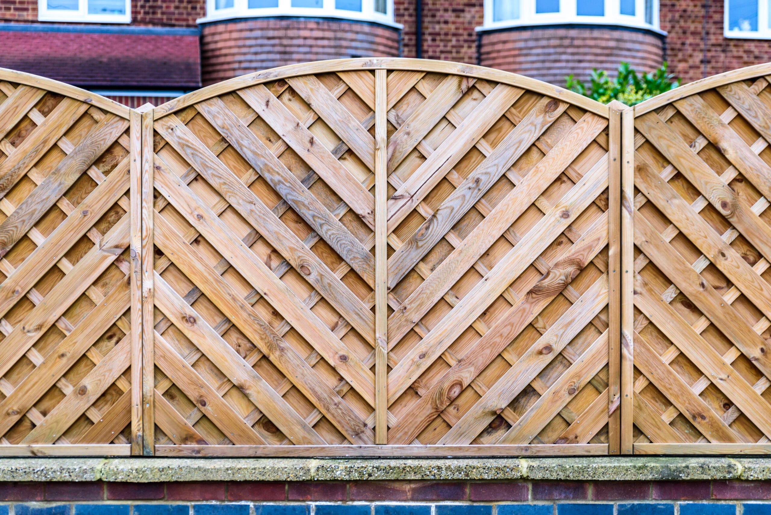 Wooden fence with a chevron pattern, featuring interwoven slats, stands on a brick base. The background shows a red-brick building with white-framed windows. The fence has a slightly arched top, adding a decorative touch.