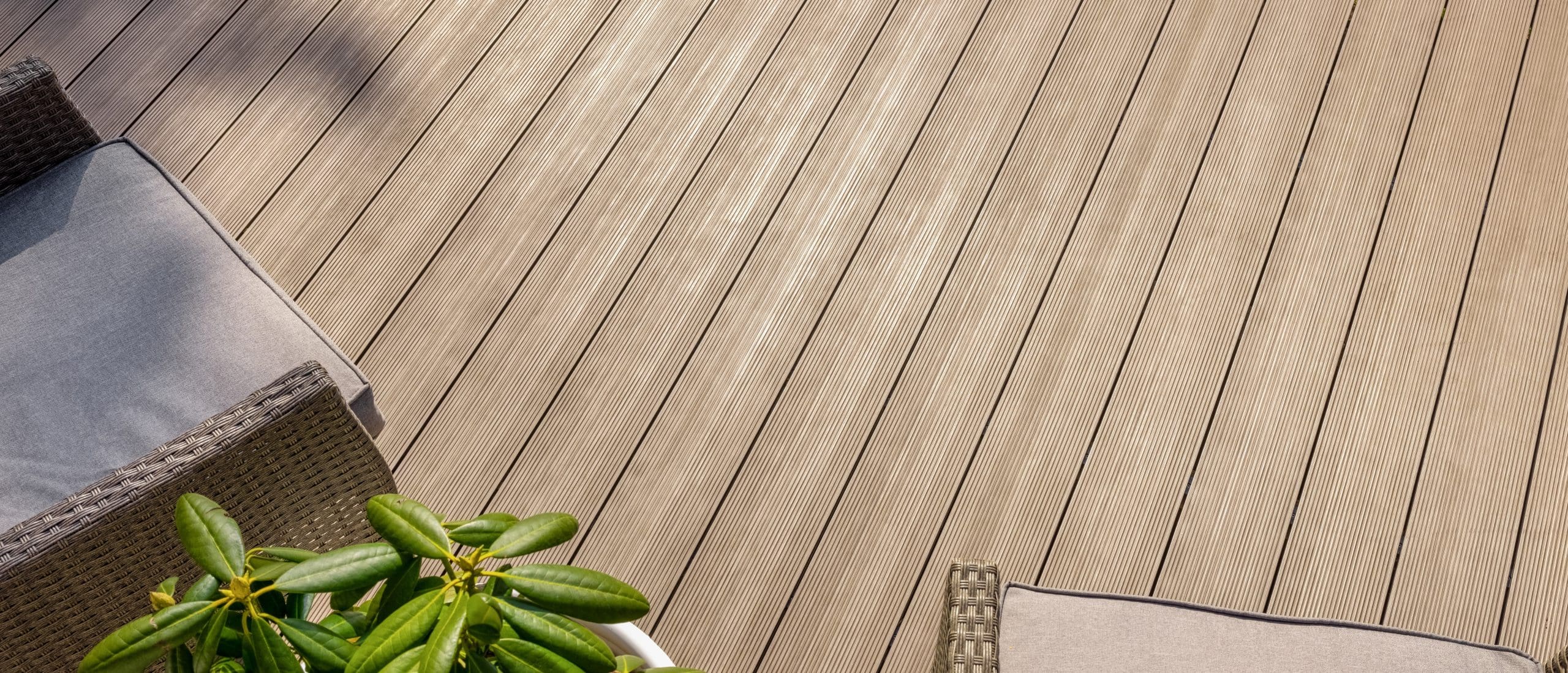 A top-down view of a wooden deck with wicker furniture. A plant with broad green leaves is in the corner of the image. The warm-toned deck has parallel planks, and cushions with neutral tones are placed on the chairs.