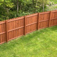 A wooden fence with a reddish-brown finish runs along a green, well-maintained grassy yard. Behind the fence, a backdrop of lush green trees and vegetation is visible. The scene is tranquil and captures a suburban or rural setting.