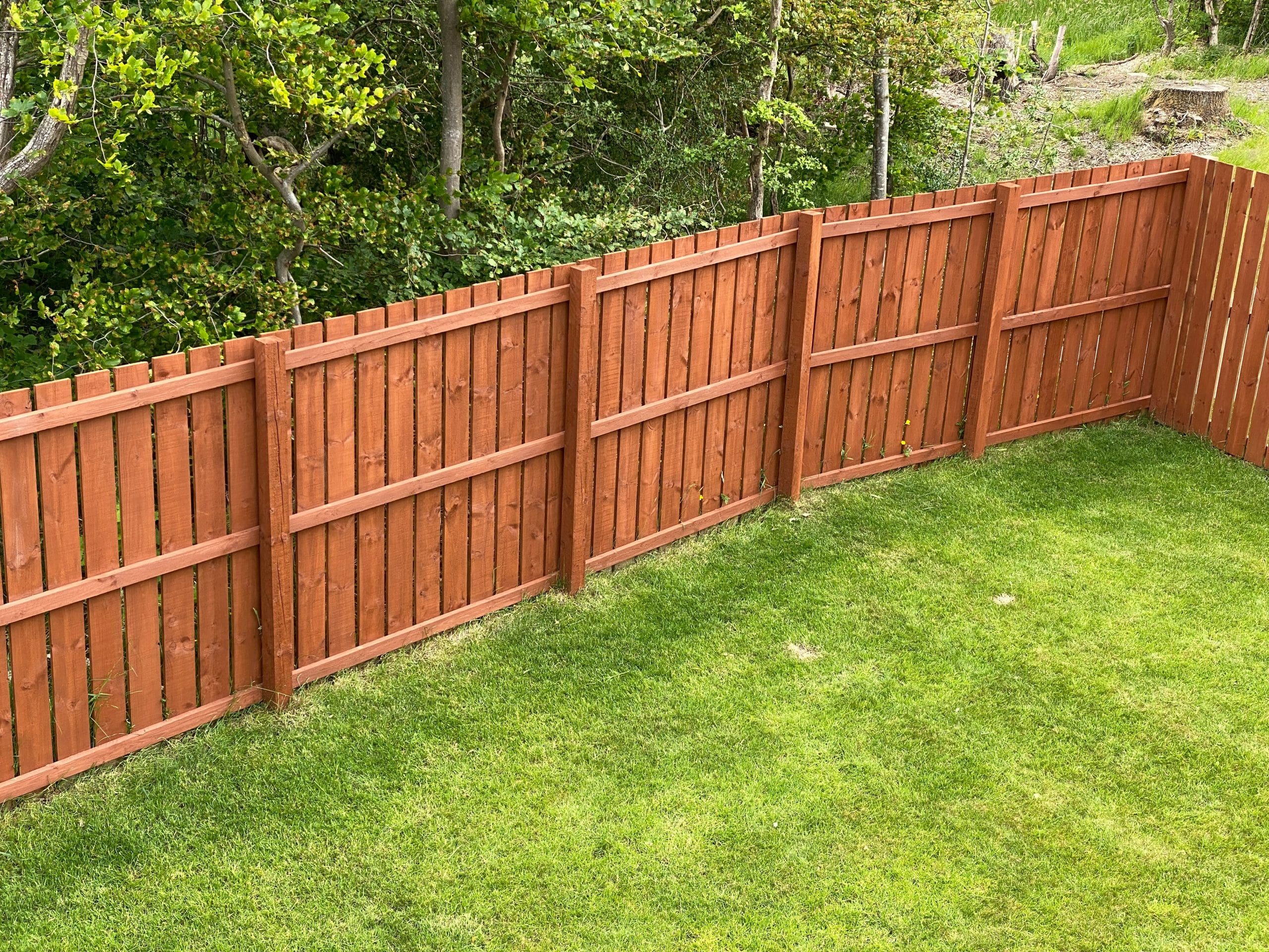 A wooden fence with a reddish-brown finish runs along a green, well-maintained grassy yard. Behind the fence, a backdrop of lush green trees and vegetation is visible. The scene is tranquil and captures a suburban or rural setting.