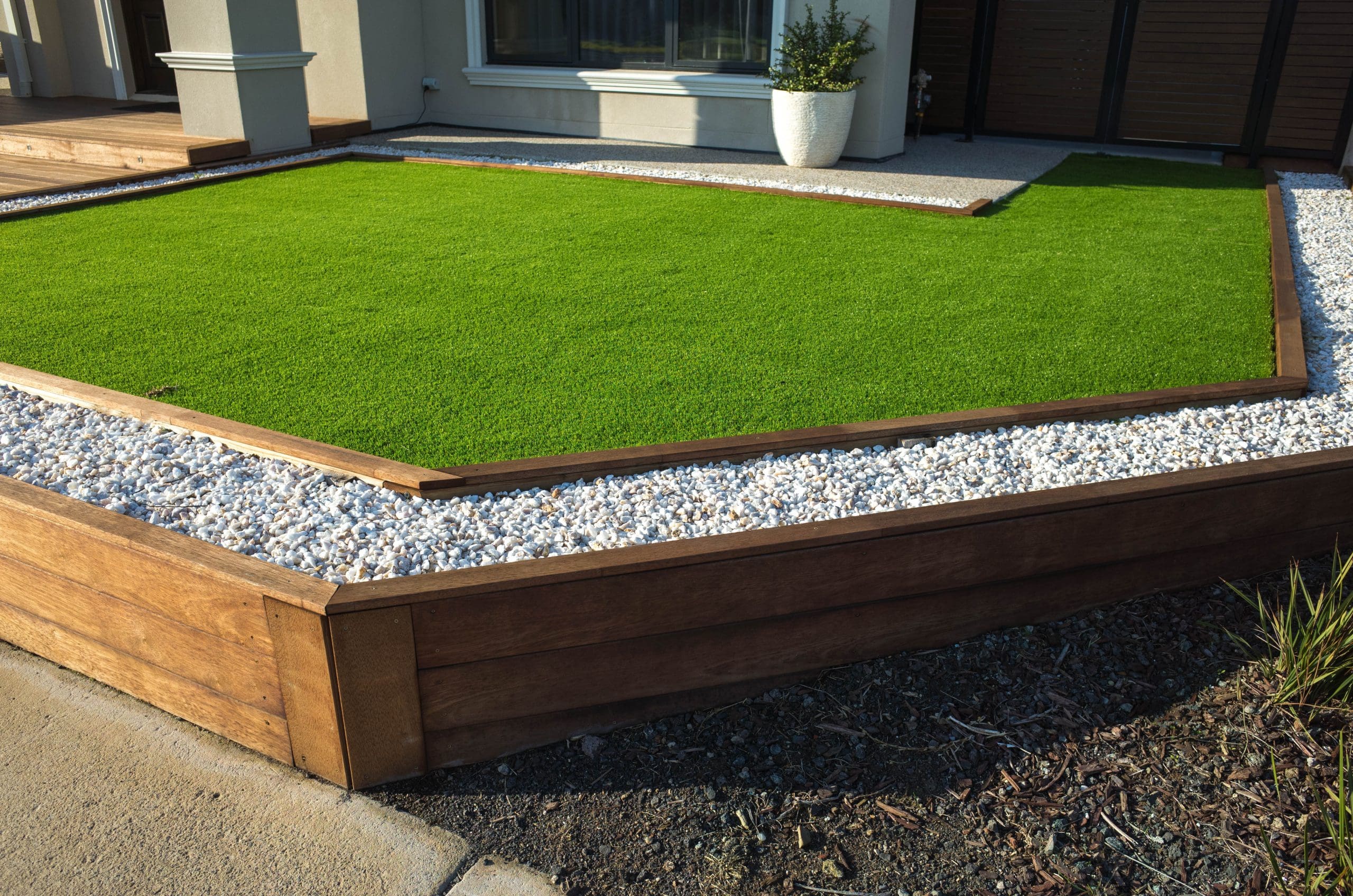 A modern backyard landscape featuring artificial grass in a geometric shape, bordered by white pebbles and wooden edges. A potted plant sits in the background near a house wall.