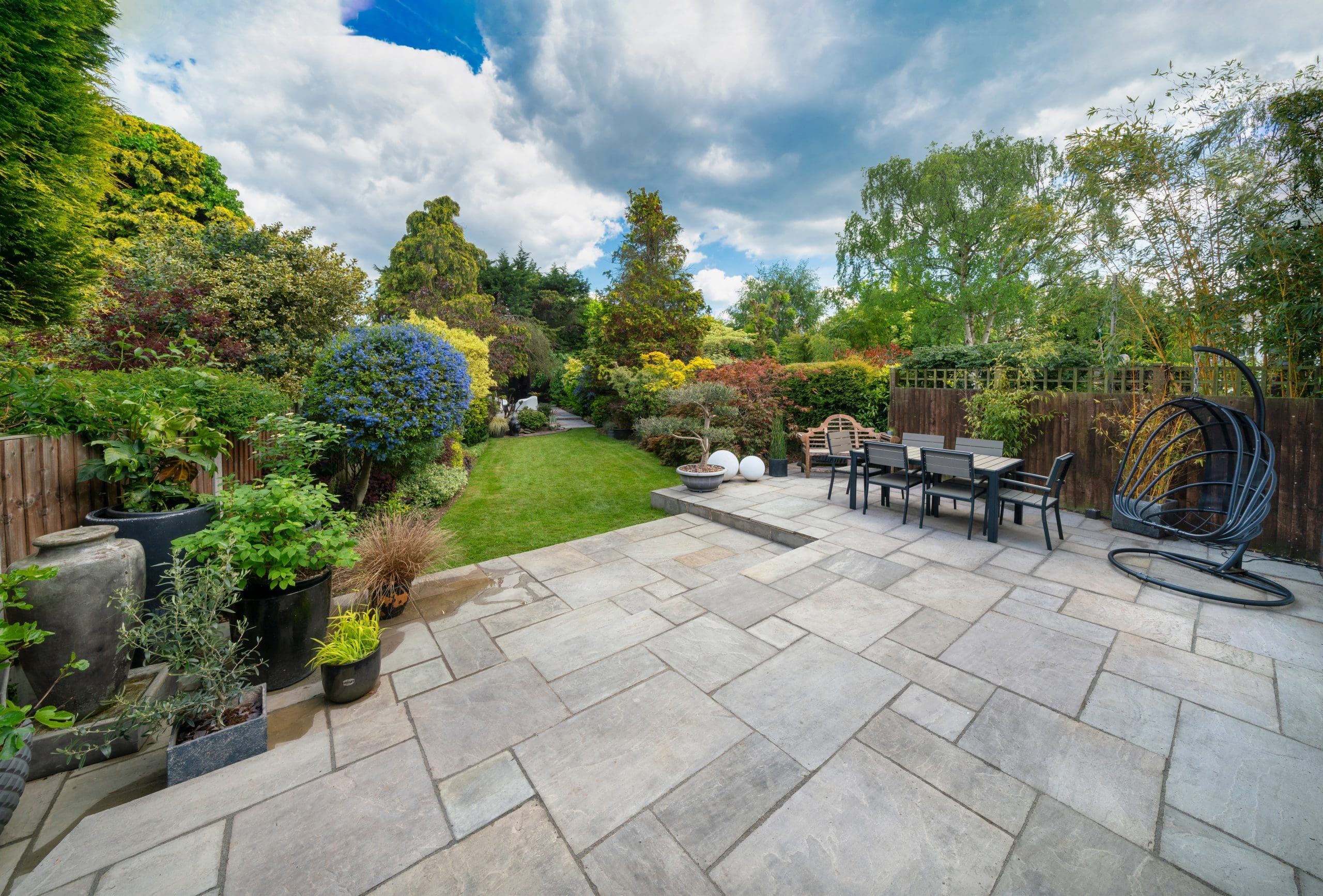 A spacious backyard with a large patio, featuring outdoor furniture and hanging chairs. The patio overlooks a lush garden with green shrubs, colorful flowers, and a well-manicured lawn, under a partly cloudy sky.