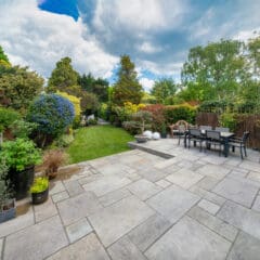 A spacious backyard features a stone patio with a dining table and chairs. The garden is lush with various trees, shrubs, and plants. The sky is partly cloudy, adding a serene ambiance to the vibrant greenery.