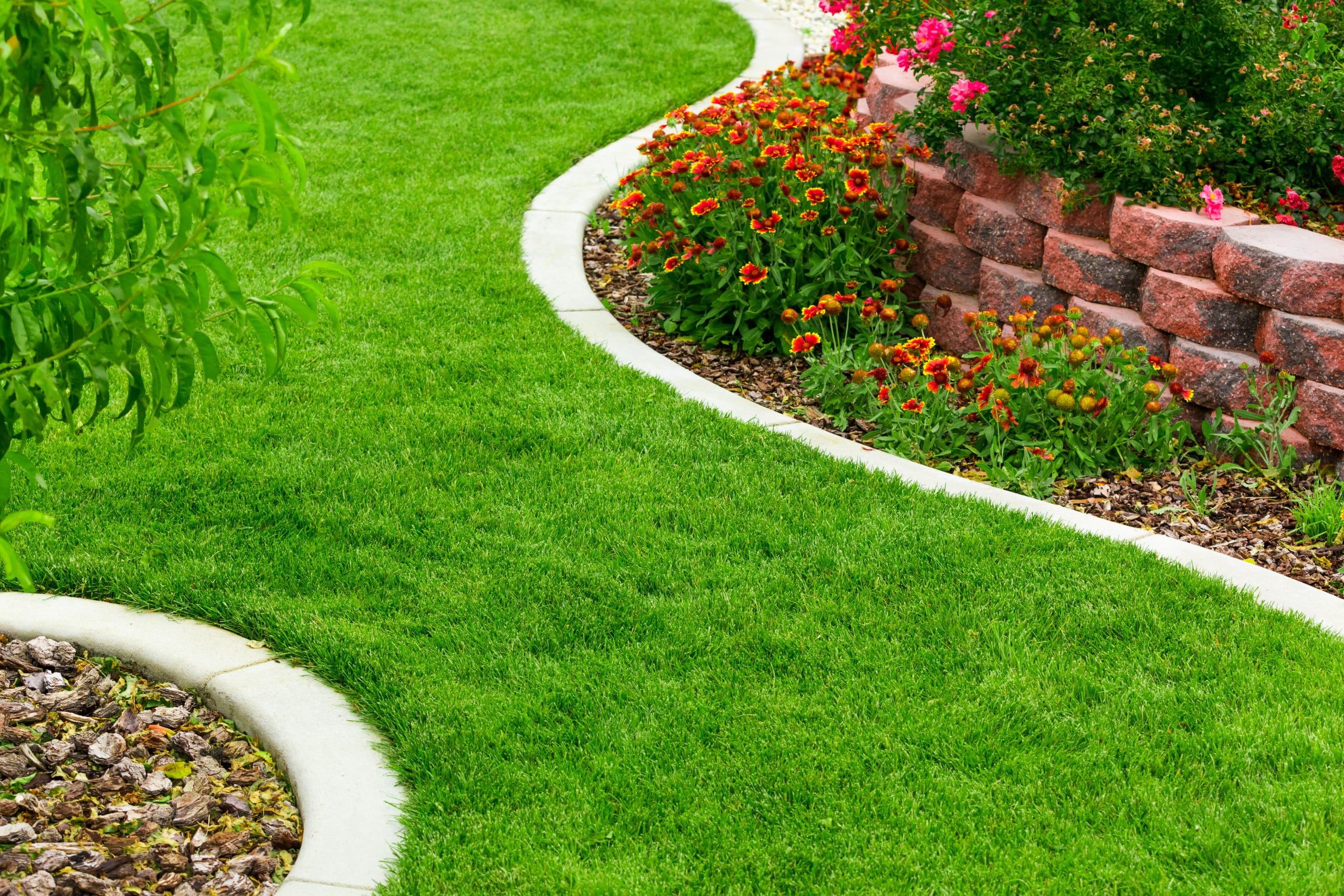 A well-maintained garden features a lush green lawn with curved stone borders. On one side, vibrant orange and red flowers bloom near a raised brick flower bed, adding a touch of color and charm to the landscape.