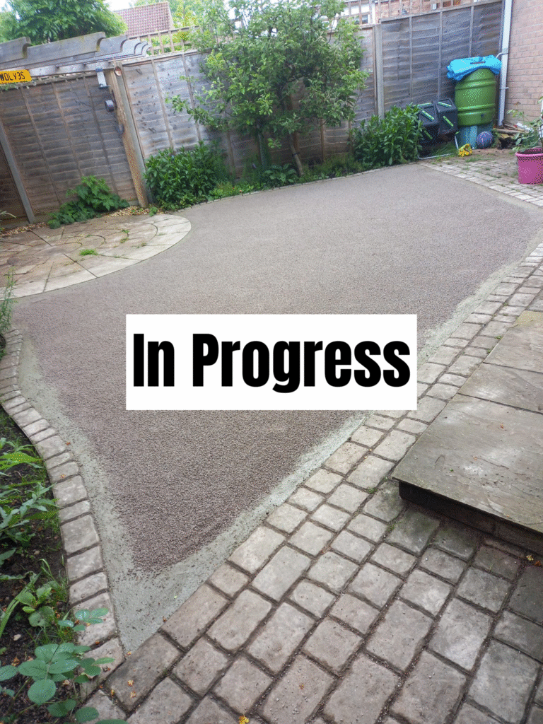 A backyard patio renovation is underway, with new stonework partially installed on the ground. The area is bordered by fencing, plants, and garden tools. A bold "In Progress" sign is overlaid on the image.