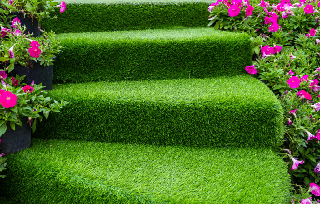 Green steps covered in artificial grass, surrounded by vibrant pink flowers and greenery on both sides, creating a lush and colorful garden scene.