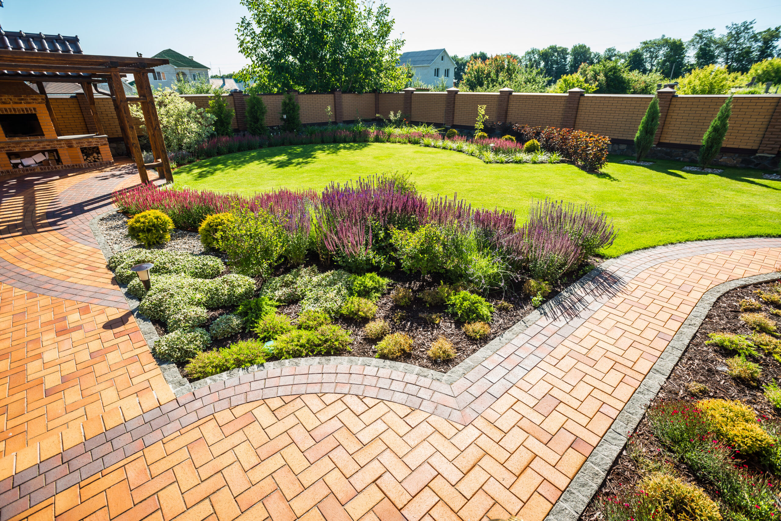 A neatly landscaped backyard with a curved brick walkway, vibrant flower beds, green grass, a wooden pergola, and a tall brick fence enclosing the space. Trees and houses are visible in the background.