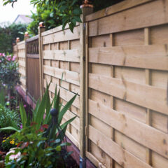 A close-up of a wooden fence beside a garden with various green plants and colorful flowers, lining a walkway in a residential area.