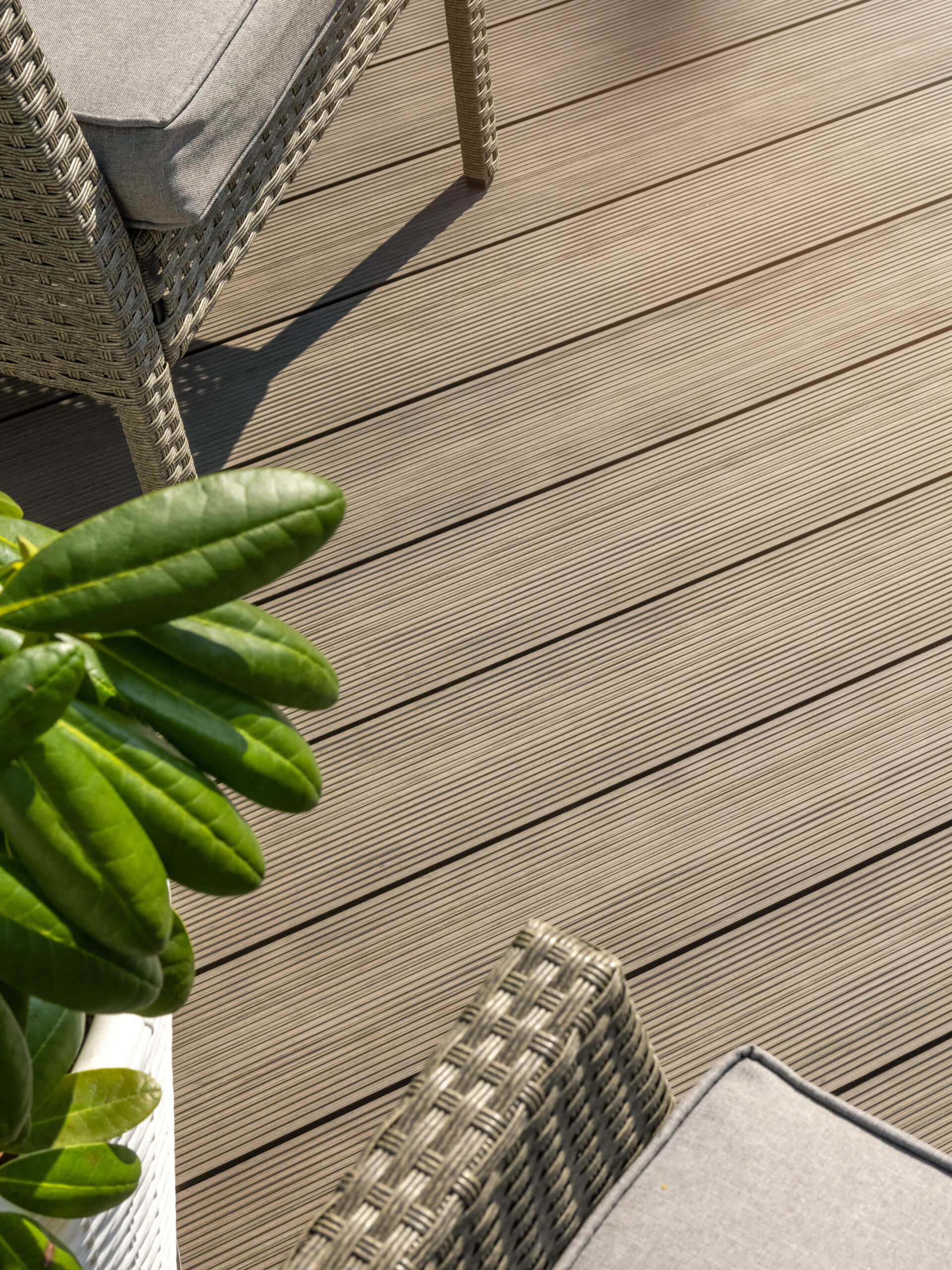 Close-up of a patio with light brown, grooved decking, part of a green leafy plant in the corner, and two wicker chairs with grey cushions partially visible in the background.