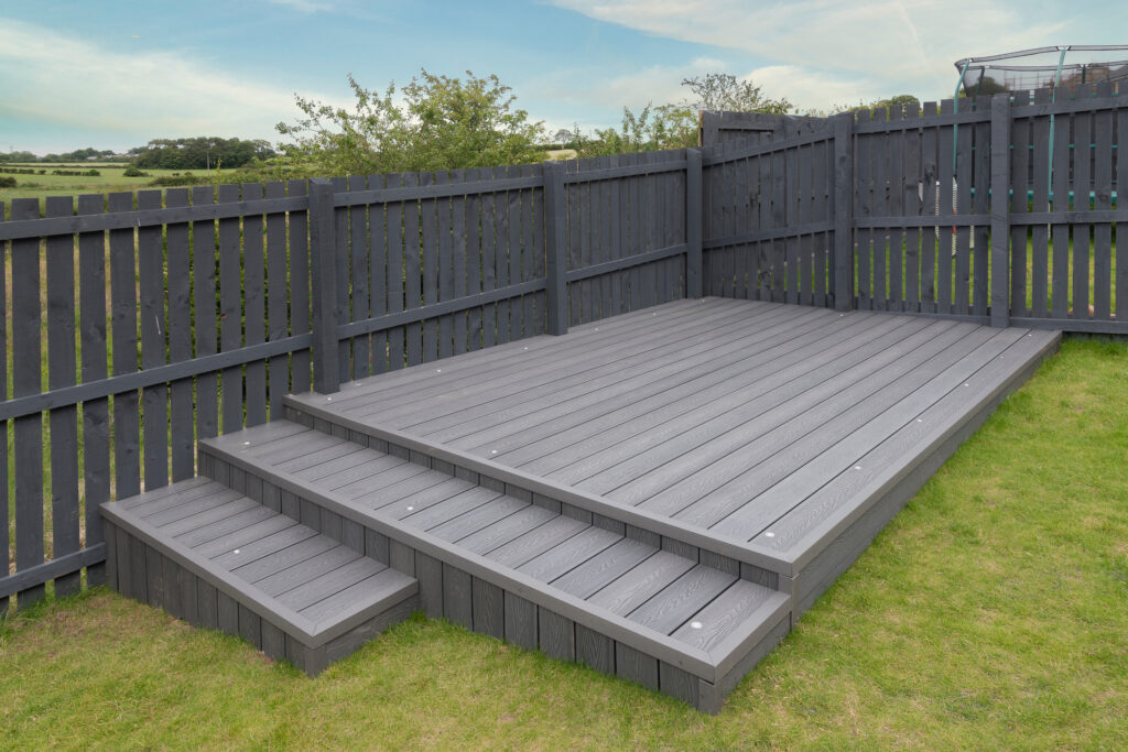 A modern backyard with a raised gray wooden deck, surrounded by a matching gray fence. Three steps lead up to the deck, and the area is set on a grassy lawn under a partly cloudy sky.