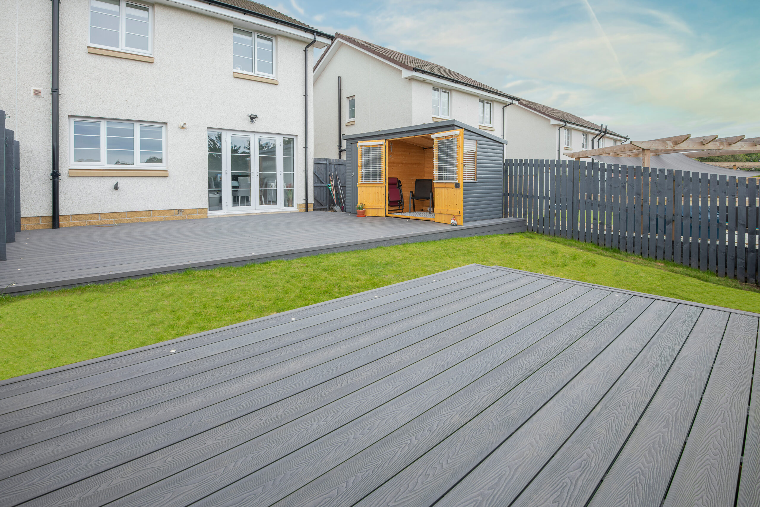 Modern backyard with gray composite decking, a green lawn, a wooden shed with seating, and a black fence, attached to a two-story white house under a blue sky with light clouds.