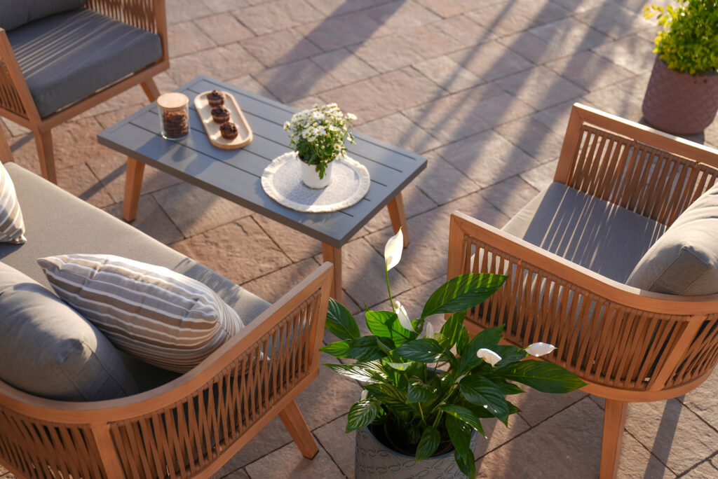 Outdoor patio seating area with wooden chairs and gray cushions around a gray coffee table, decorated with a small flower arrangement and candles. A potted green plant and striped cushion are also visible.