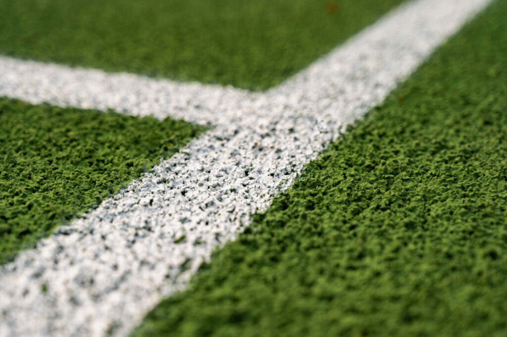 Close-up view of a textured green sports field with a sharp, white painted line marking, likely from a tennis or soccer court, captured at an angle for a detailed and dynamic perspective.