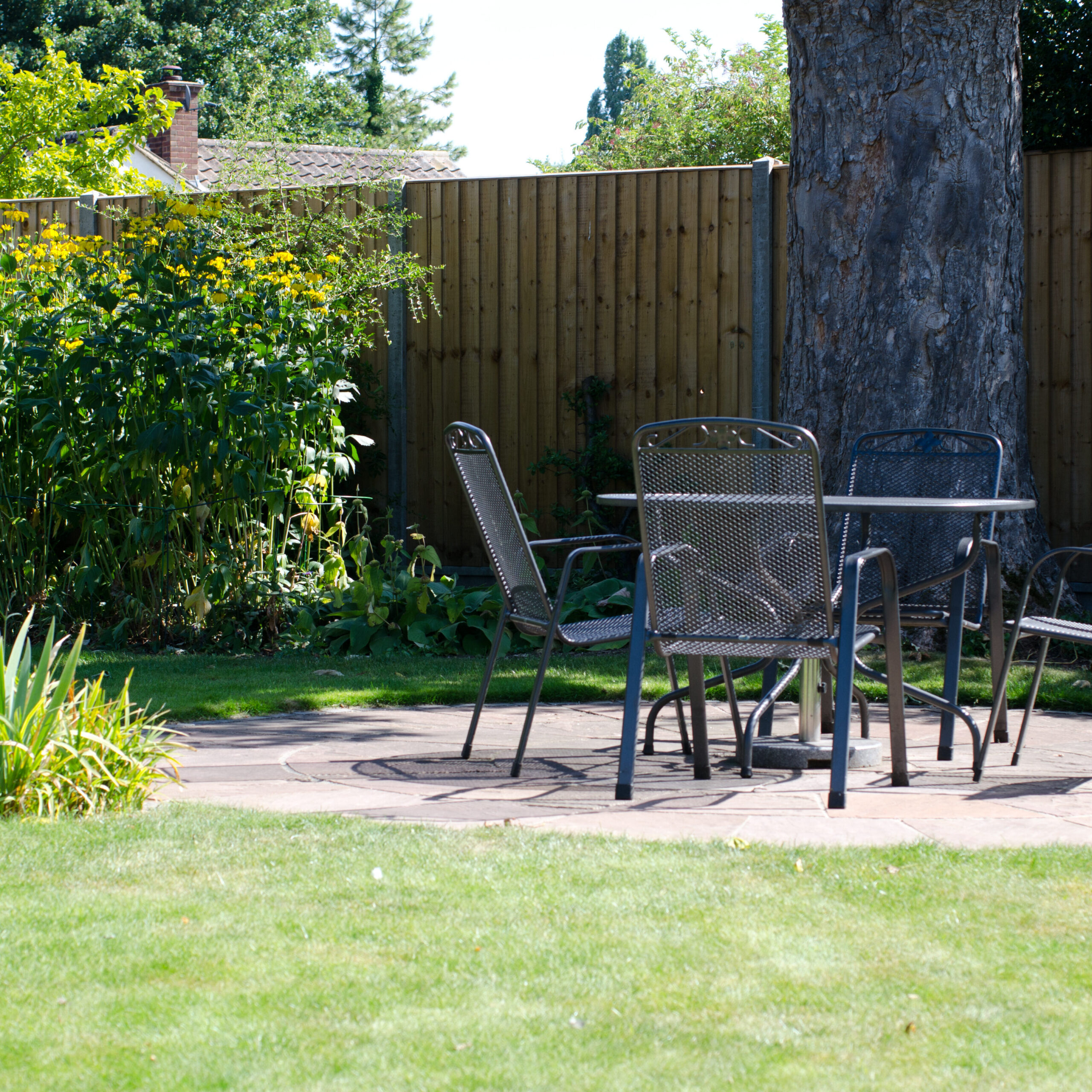 A patio area in a sunny garden with a round metal table and four matching chairs. The patio is surrounded by green grass, shrubs, and a tall wooden fence with a large tree nearby.