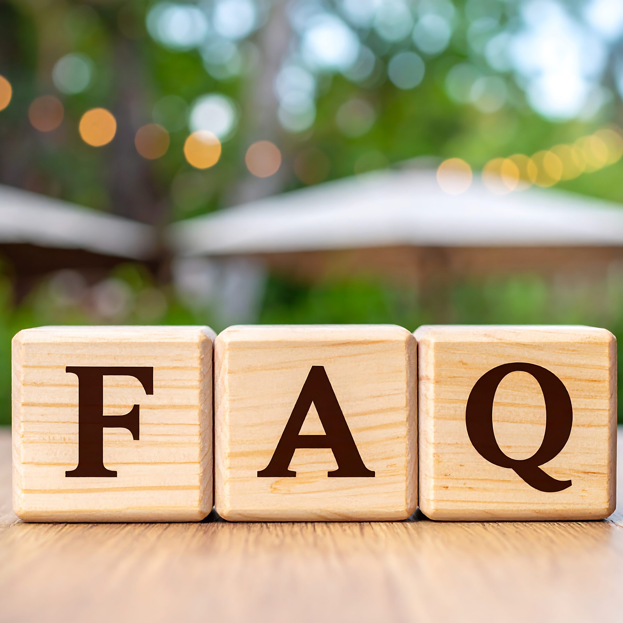Three wooden blocks with the letters "F", "A", and "Q" are arranged in a row on a wooden surface. The background is blurred greenery with bokeh lights.