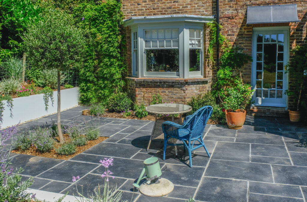 A sunny garden patio with slate tiles, a small round table, and a blue wicker chair. There are potted plants, a tree, flowers, and a brick house with a bay window and glass door in the background.
