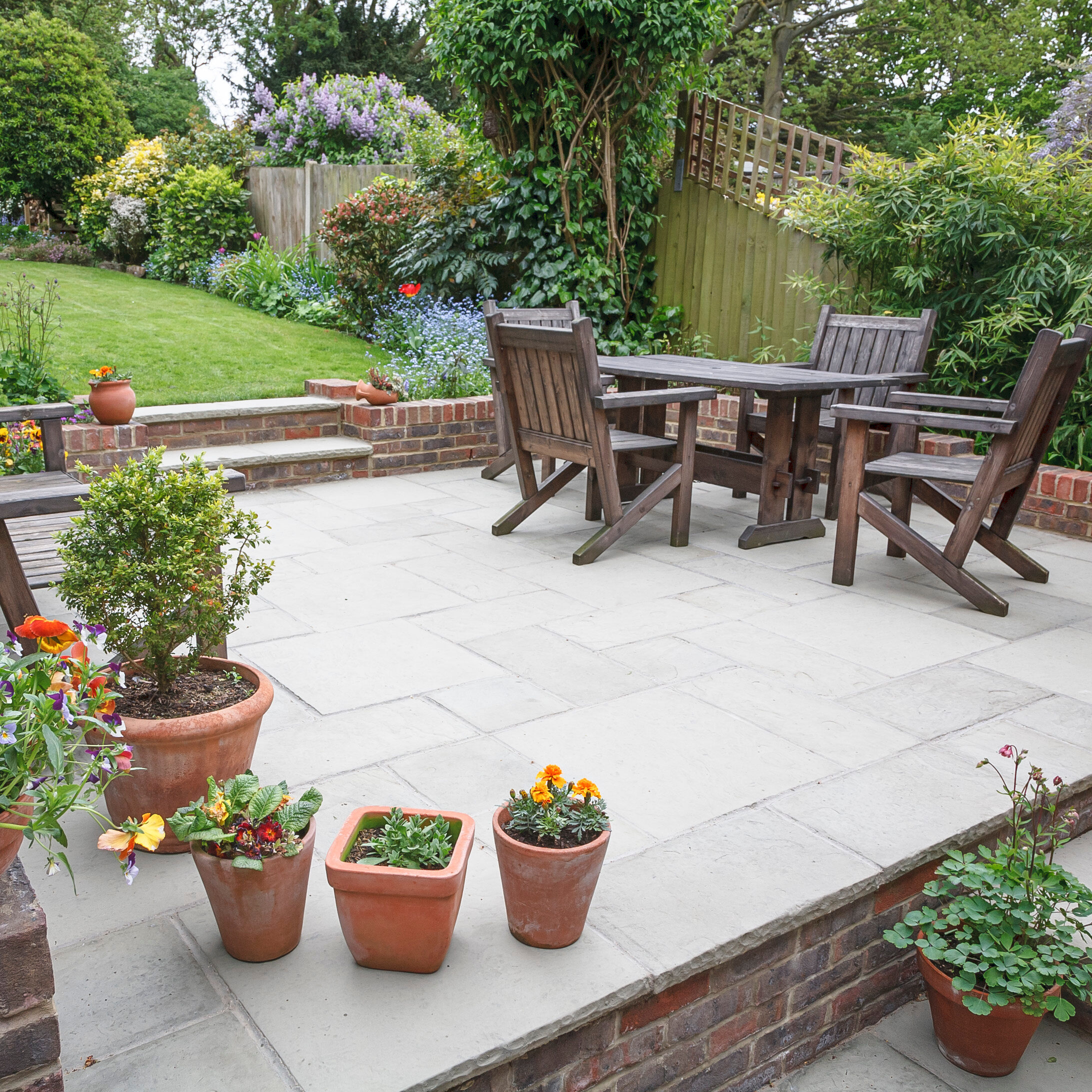 A patio with wooden table and chairs sits next to a grassy garden, surrounded by plants, flowers, and potted plants on a stone floor. A wooden fence and lush greenery are in the background.