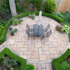Aerial view of a circular garden patio with a square table, six chairs, and a closed umbrella, surrounded by neatly trimmed hedges, flowering plants, and a green lawn.