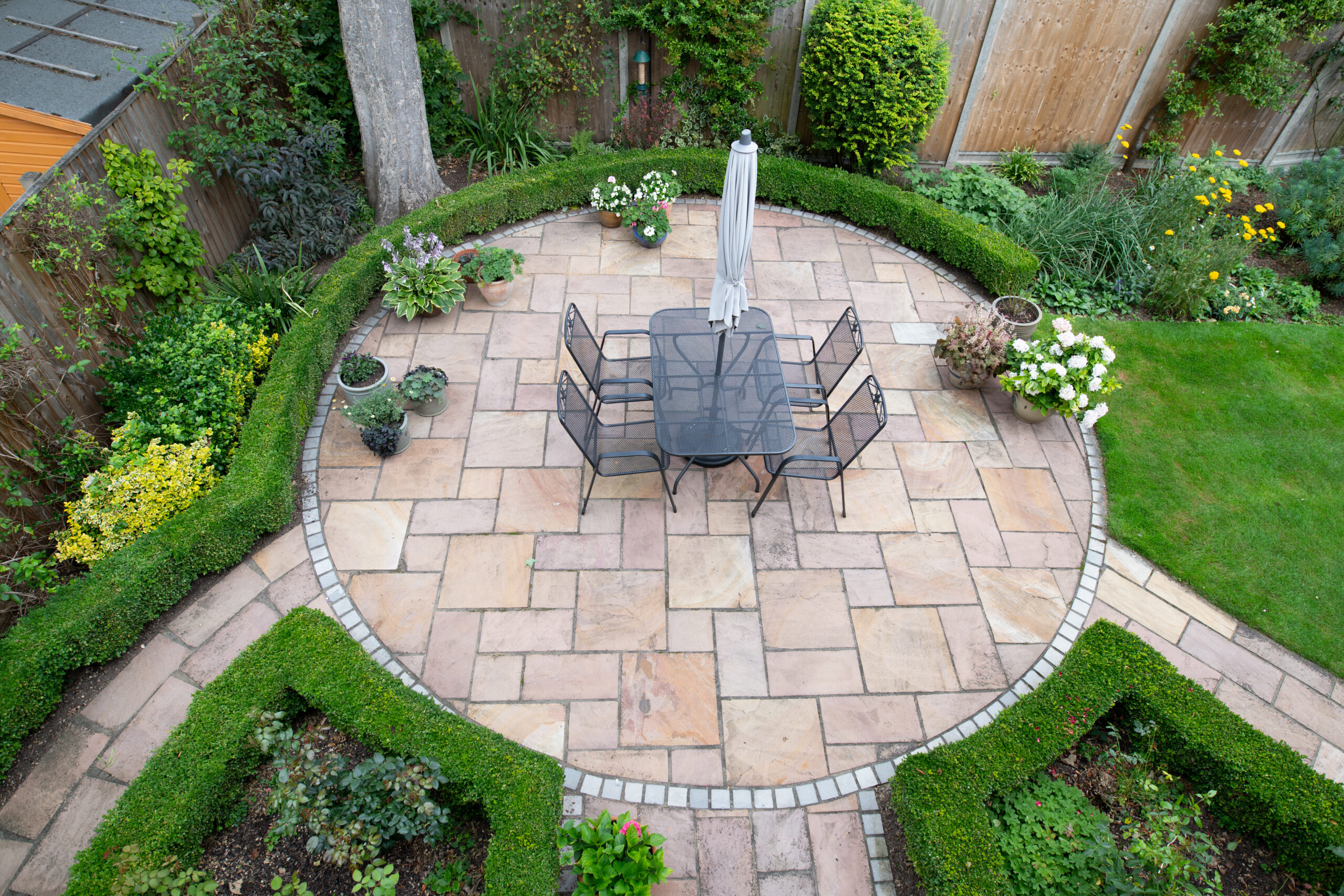 Aerial view of a circular garden patio with a square table, six chairs, and a closed umbrella, surrounded by neatly trimmed hedges, flowering plants, and a green lawn.